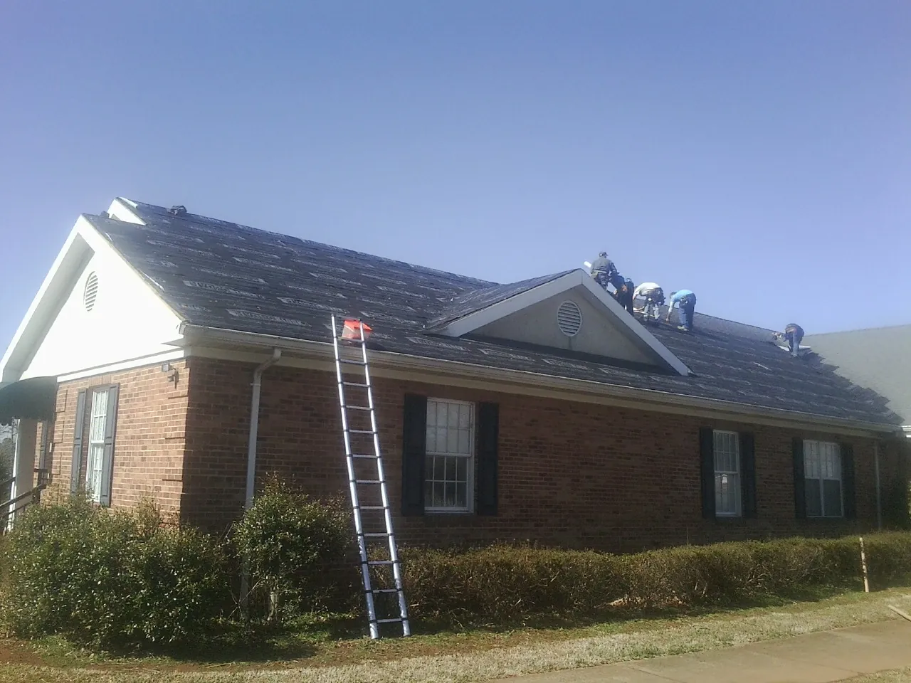Crew tearing shingles off a brick ranch roof with a ladder set — Emergency Roof Repair work by our crew in Keego Harbor