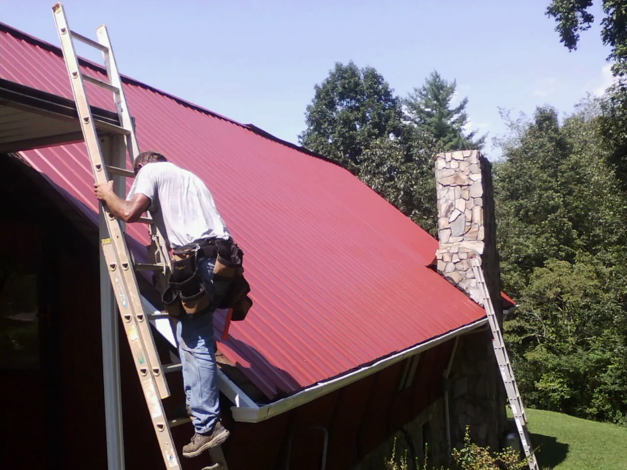 Roofer on a ladder beside a red metal roof and stone chimney — Soffit & Fascia Repair by Roofing Brothers in Keego Harbor